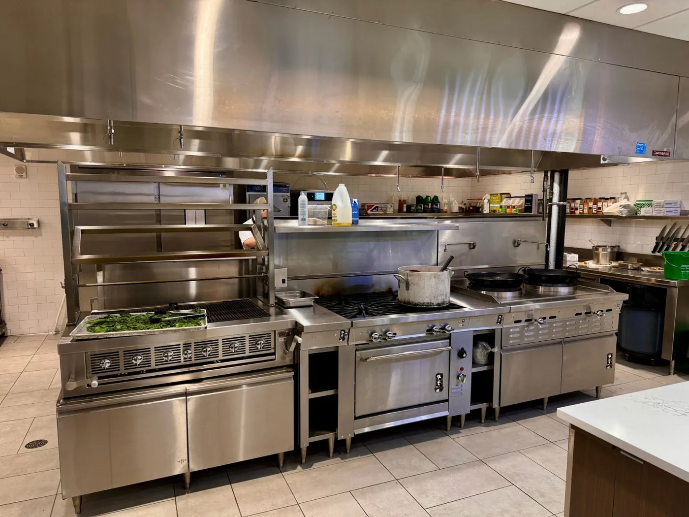 A stainless steel commercial kitchen cook line with range, griddle, charbroiler, and exhaust hood